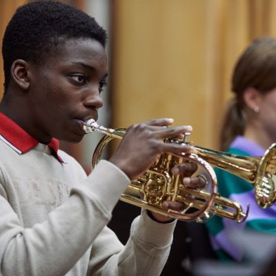 A boy wearing a white jumper, with a red collar sticking out of it, plays the trumpet
